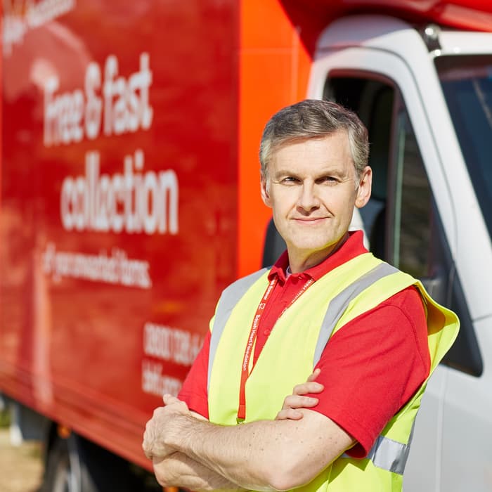 Man in a high-visibility vest stands confidently with crossed arms in front of a red British Heart Foundation truck labeled "Free & fast collection," outdoors near a building.