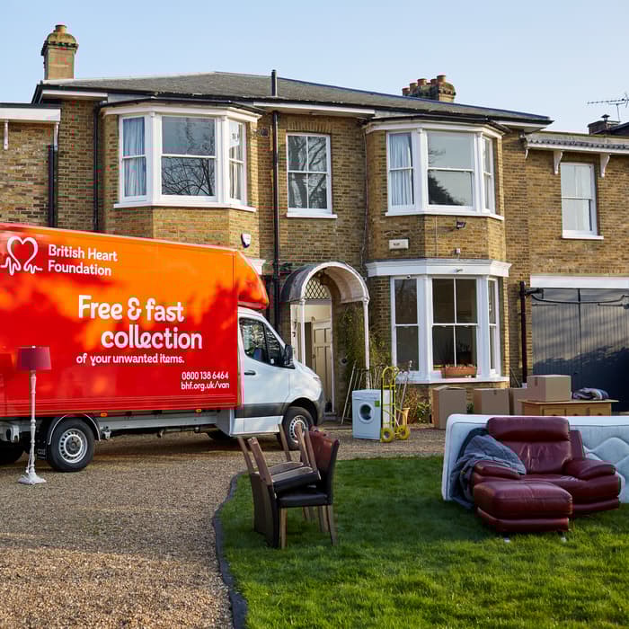 A red British Heart Foundation truck parked outside a suburban house, collecting furniture, including a sofa and chair, with sign reading "Free & fast collection of your unwanted items.”