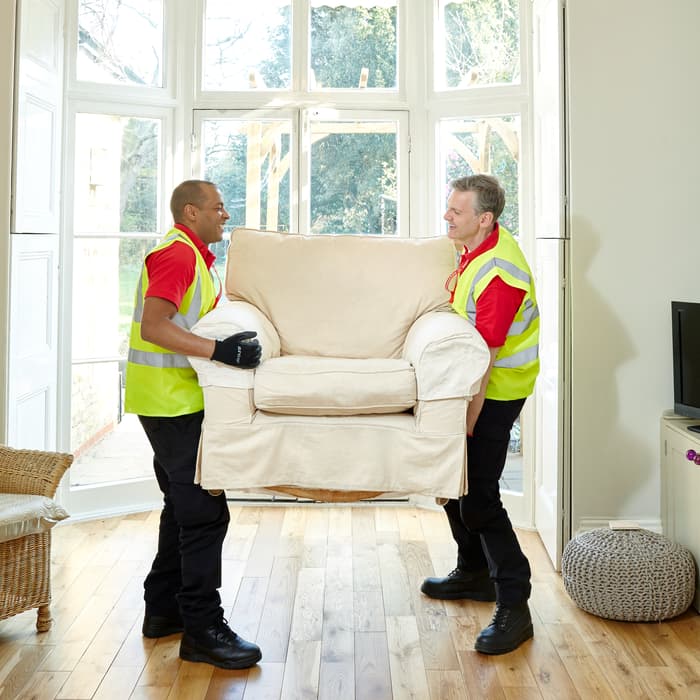 Two men wearing yellow safety vests lift a beige armchair in a bright room with wooden flooring, large bay windows, and wicker furniture.