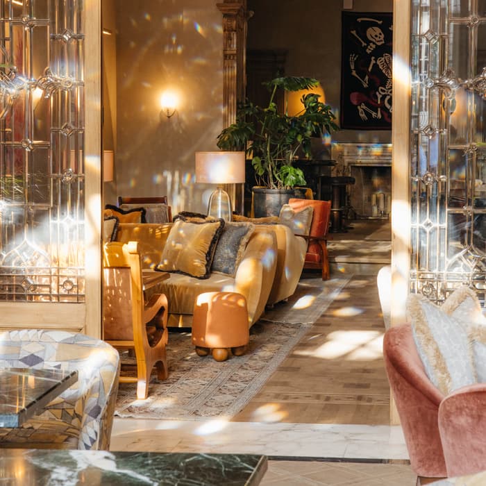 Luxurious living room features plush sofas, ornate lamps, and lush plants. Elegant stained-glass doors and chandeliers enhance the sophisticated ambiance. Sunlight filters through the room, creating a warm, inviting atmosphere.