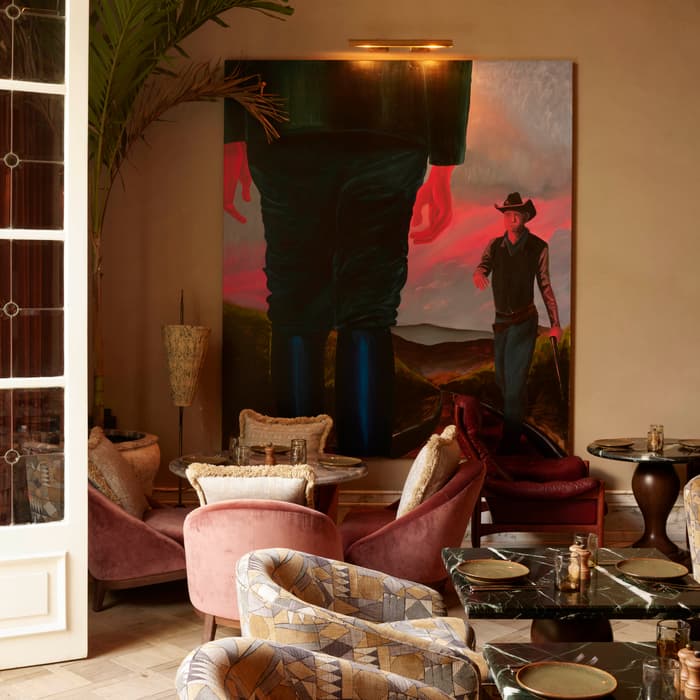 Large painting of a figure in a hat dominates a cozy, tastefully-decorated café with plush chairs, green marble tables, and warm lighting. Palm fronds and open French doors add elegance.