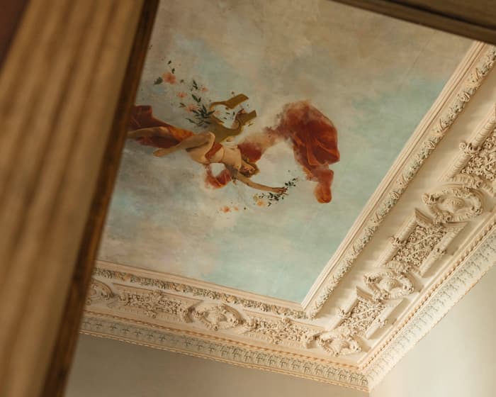 A fresco painting of a figure holding flowers adorns an ornate, intricately detailed ceiling inside a classical, elegant interior with decorative molding.