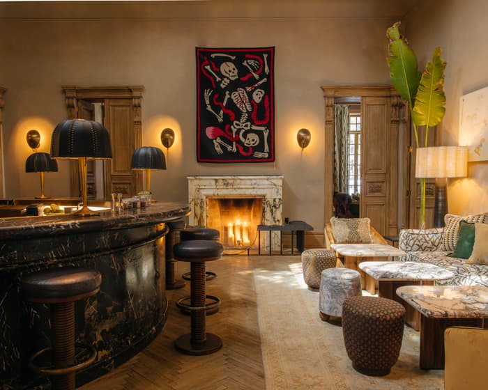 A cozy, dimly lit room featuring a marble bar counter with bar stools, a plush seating area with patterned cushions and tables, and a lit fireplace beneath an eclectic artwork of skeletons.