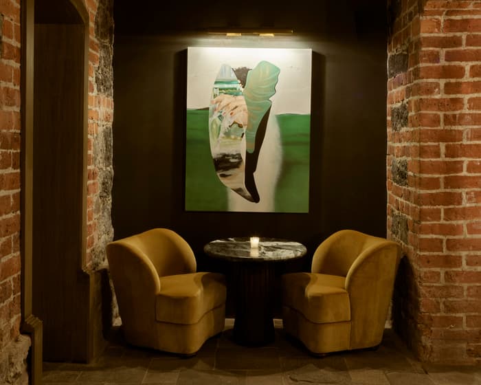 Two yellow velvet chairs and a round black marble table sit against a painted wall with a large abstract artwork, framed by exposed red brick archways and dim lighting.