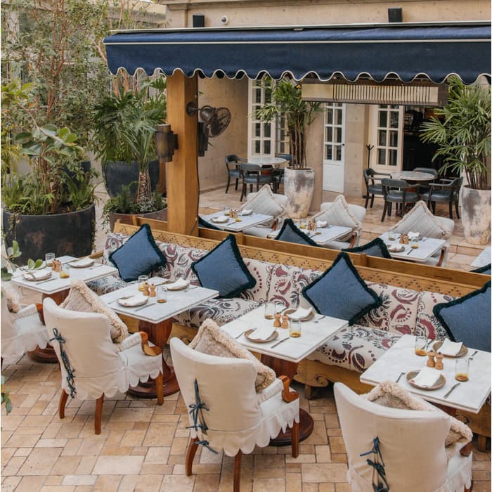 Outdoor dining area with cushioned chairs and blue pillows arranged around tables beneath a blue awning. Surrounded by potted plants and overlooked by a beige, columned building with balconies.