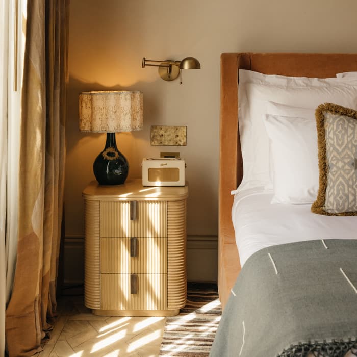 A wooden nightstand with a lamp, a small beige radio, stands beside a neatly made bed with white linens and a gray blanket, in a warmly lit bedroom. Shadows from window blinds fall across the floor.