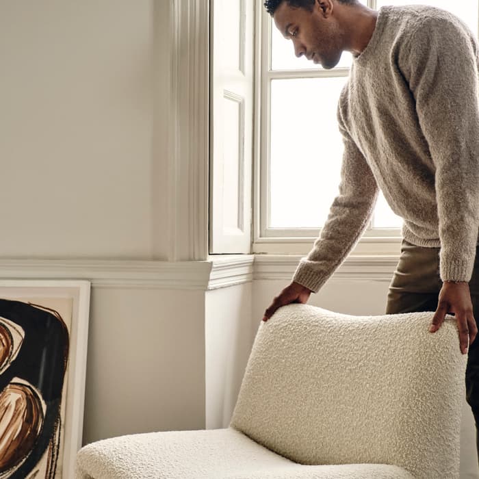 A man, dressed in a beige sweater, adjusts a modern, textured, cream-colored chair beside a sunny window. A framed abstract painting leans against the wall on a patterned rug.