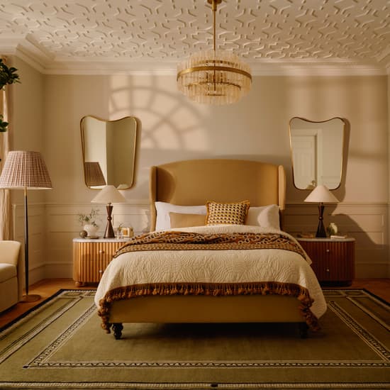 A bed with a beige headboard and decorative pillows is situated in a stylish bedroom, featuring matching nightstands, mirrored walls, a chandelier, an armchair, and a textured ceiling.