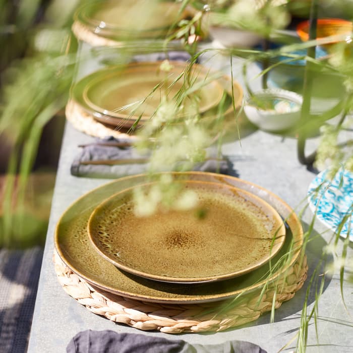 Green ceramic plates are stacked on wicker placemats with gray napkins and silverware beside them on an outdoor concrete table, surrounded by tall grass and other tableware.