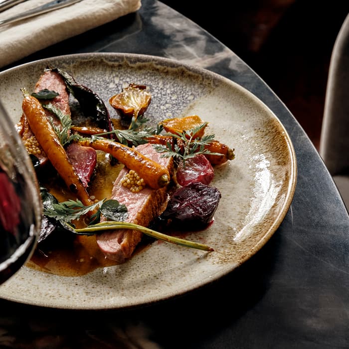 A plated dish featuring seared meat and various roasted vegetables on a ceramic plate, placed on a dark marbled table near a folded napkin and cutlery.