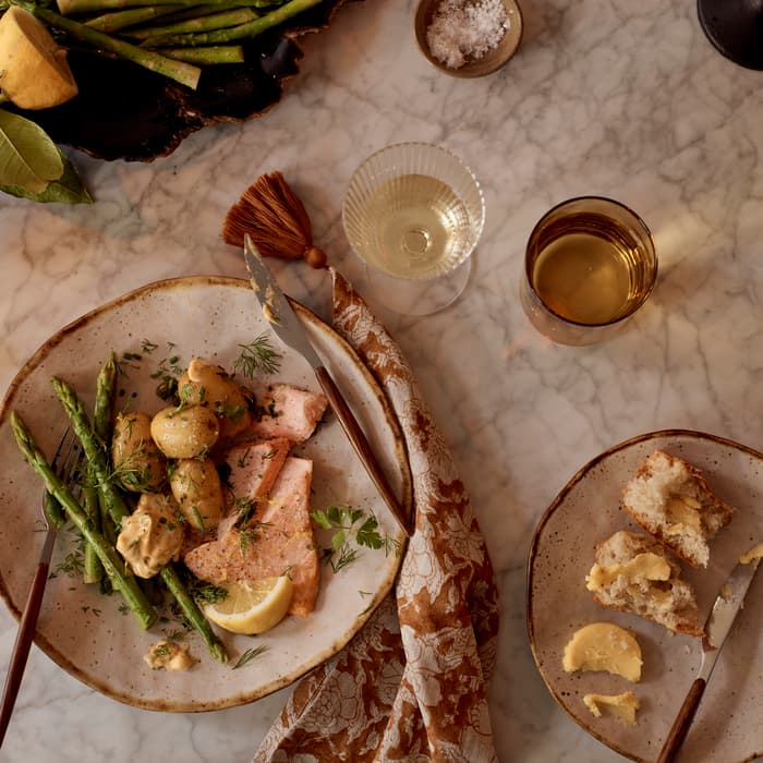 A dinner scene with a main plate of salmon, potatoes, and asparagus, accompanied by a small plate with bread and butter on a marble table, alongside two beverages and seasoning.