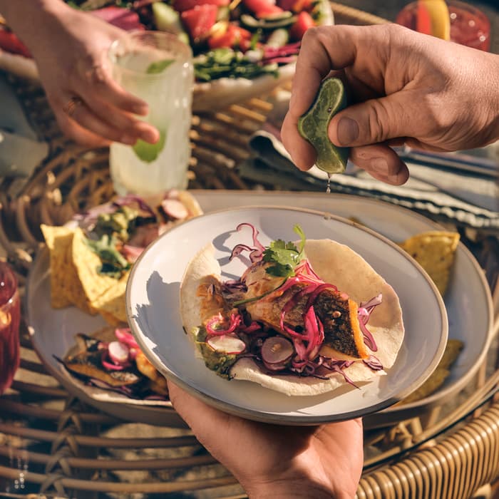 A hand holds a plate with a taco while another hand squeezes lime juice over it. Surrounding wicker table has nachos, a drink, and a bowl of salad.