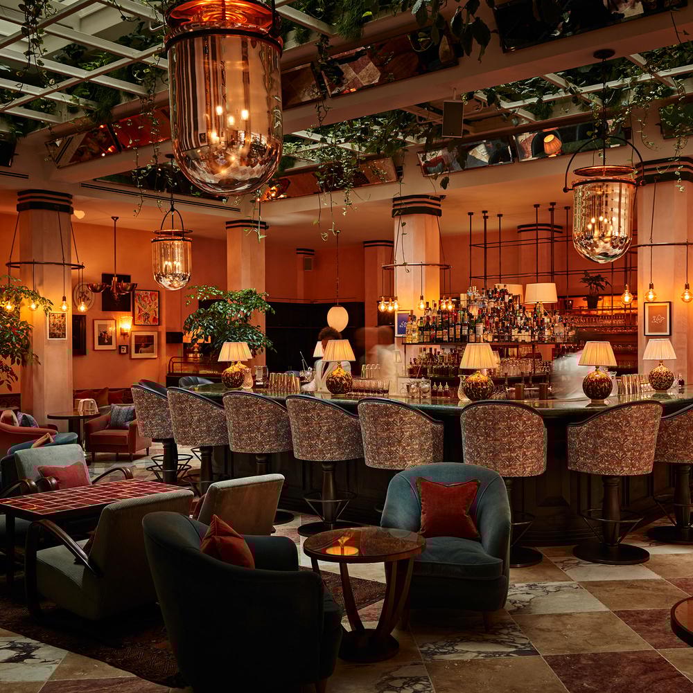 Luxurious bar with upholstered stools, softly glowing hanging lights, and plants in an upscale lounge setting featuring cozy armchairs, patterned flooring, and framed artwork on peach walls.