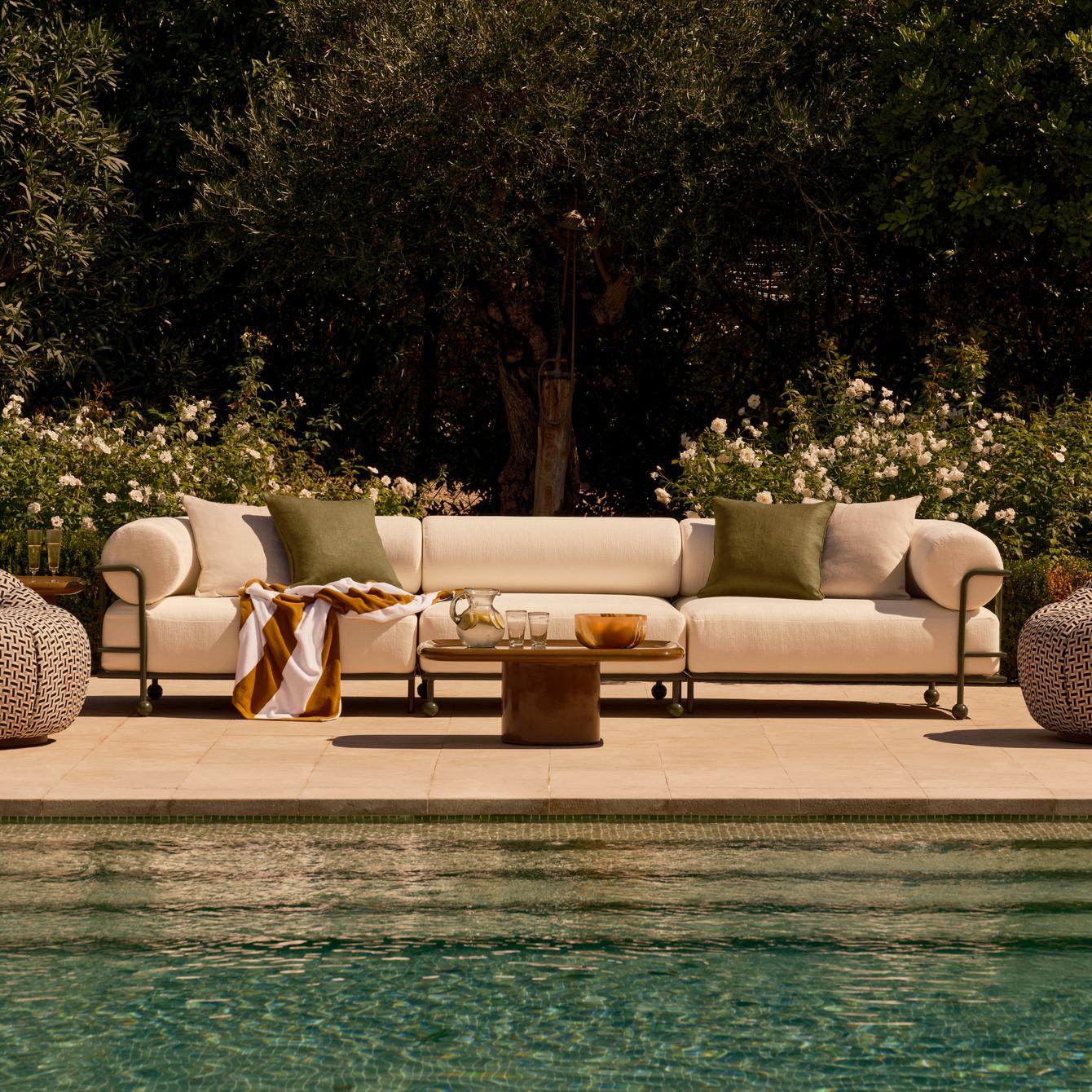 A white couch with green pillows and a striped blanket rests by a poolside, with a wooden table nearby holding glasses and a bowl; surrounded by lush green bushes and trees.