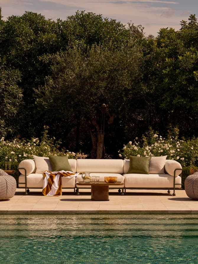 A white couch with green pillows and a striped blanket rests by a poolside, with a wooden table nearby holding glasses and a bowl; surrounded by lush green bushes and trees.