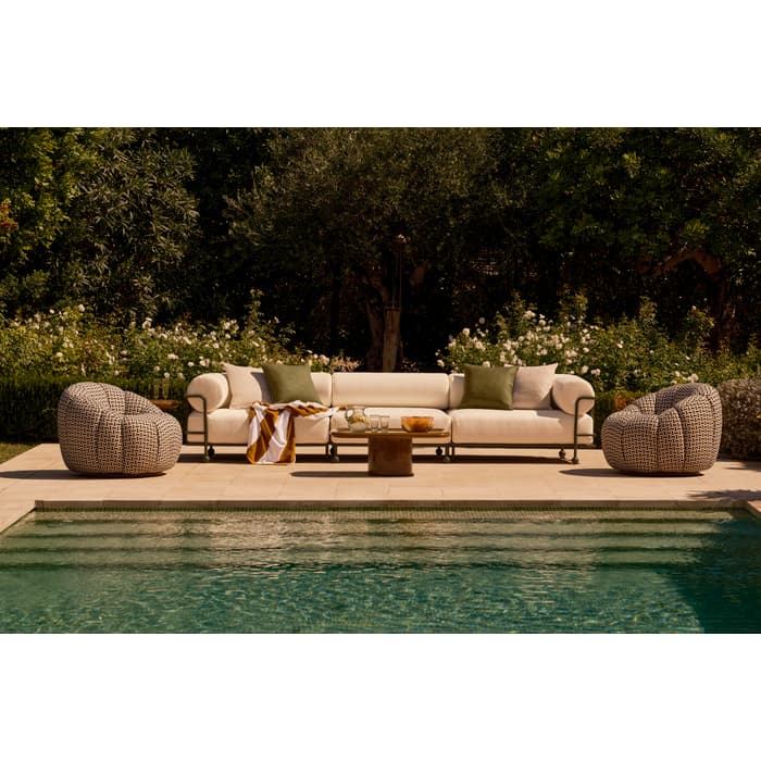 A white sofa and two patterned armchairs are positioned on a patio adjacent to a swimming pool, surrounded by lush greenery and flowering bushes.