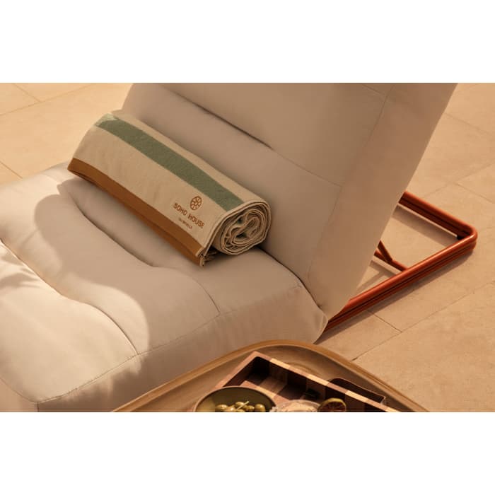 A cushioned lounge chair with a rolled-up green and white towel labeled "SOHO HOUSE" lies on a wooden patio next to a tray holding two cocktails and a bowl of olives.
