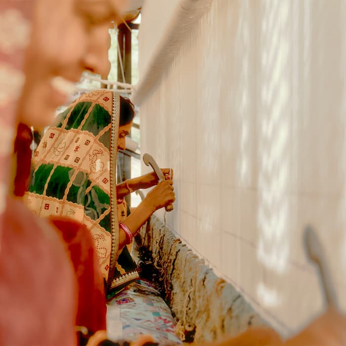 Women with colorful veils work on a large white tapestry, using small tools for embroidery, in a sunlit room.