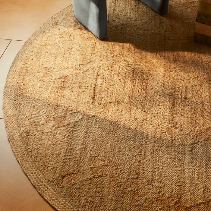 Round, woven, light-brown rug is placed on tan wood flooring. Part of a gray upholstered chair leg is visible on the rug.