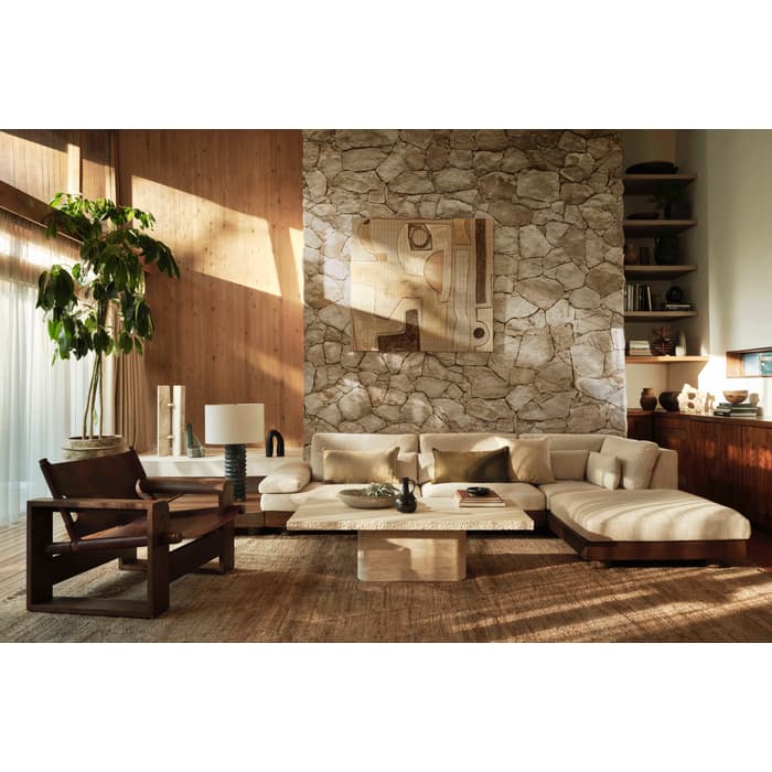 Modern living room displaying neutral-toned furniture, including a beige sectional sofa, a wooden armchair, a stone coffee table, and a floor lamp, against a stone accent wall and large potted plant by a sunlit window.