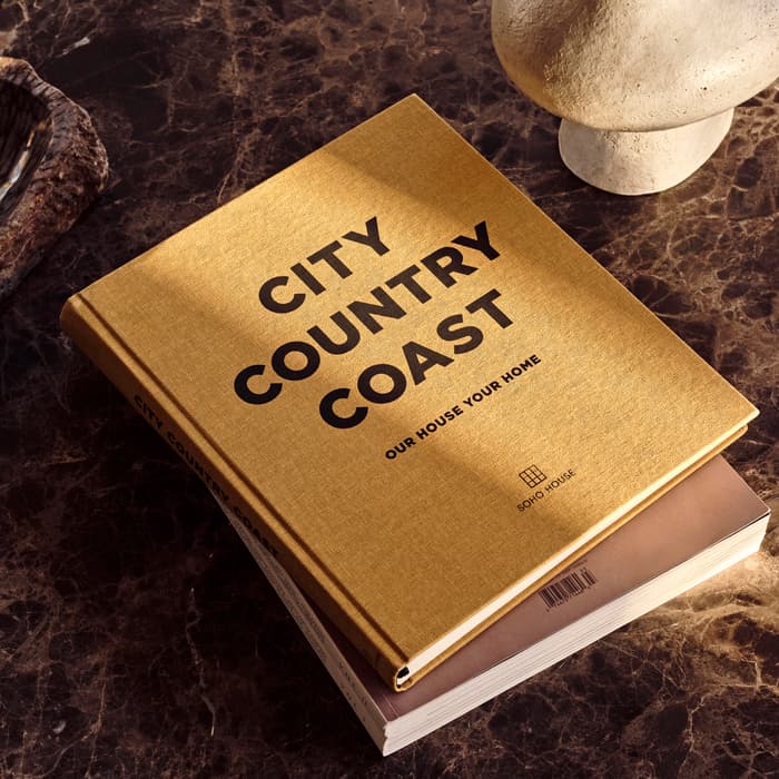 A gold-colored book titled "CITY COUNTRY COAST: OUR HOUSE YOUR HOME" rests on a dark marble table, alongside a textured bowl and a stone sculpture.