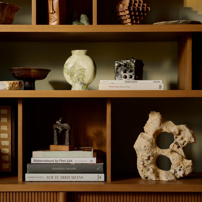 A wooden bookshelf with various decorative items: carved wooden face, pottery, abstract sculptures, and stacked books titled "Alvar Aalto," "Philip Guston," "Modern Originals," "Watercolours by Finn Juhl," "François Halard," and "Haute Bohemians."