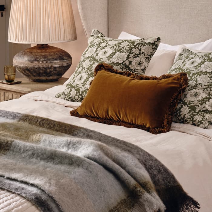 Bed with cream headboard and layered pillows, one being brown velvet, and a striped blanket; beside a wooden nightstand with a lamp and cup; situated in a cozy, warmly-lit room.