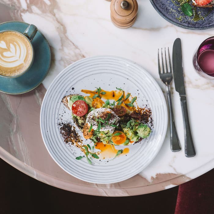 A marble table holds a latte, two plates of food (one with avocado toast, one with strawberries), utensils, wooden salt and pepper shakers, and a purple glass, above a red velvet chair.