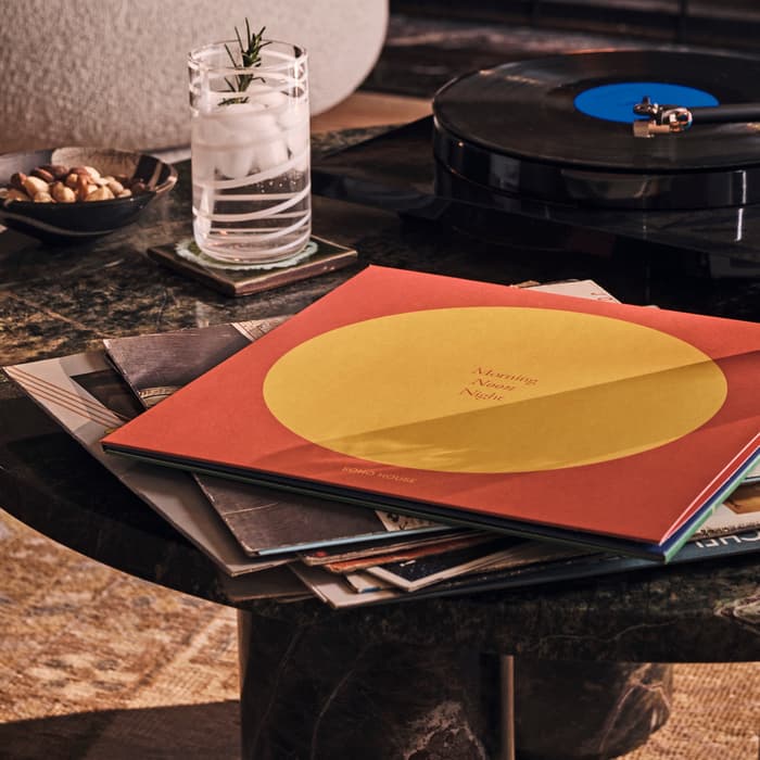 A record player spins a vinyl on a dark marble table beside a drink with rosemary and nuts. Nearby, an album titled "Morning Noon Night" rests, in a cozy, fireplace-lit room.