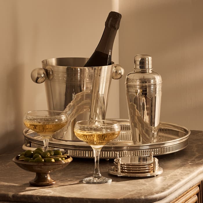 A bottle of champagne rests in a silver ice bucket on a tray beside a cocktail shaker, with two filled champagne glasses and a bowl of olives on a marble countertop.
