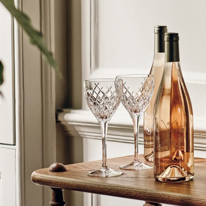 Six wine bottles and two crystal glasses are arranged neatly on a wooden table near a large window, with a potted plant and a white sculpture nearby in a softly lit room.
