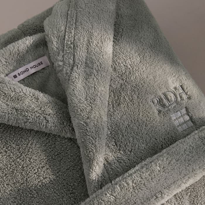 A folded, light-grey, plush bathrobe featuring the text "SOHO HOUSE" on an inner label and "RDH" with a grid square embroidered on the exterior, placed on a neutral-toned surface.