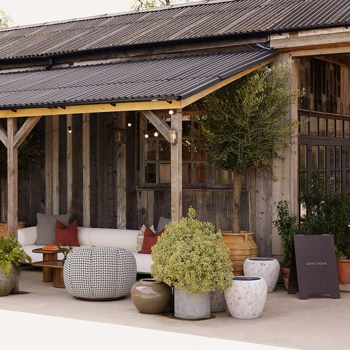 Outdoor seating area with cushioned furniture, poufs, and potted plants arranged under a rustic wooden awning, part of a wooden building with vertical planks and string lights. Sign reads "SOHO HOME."