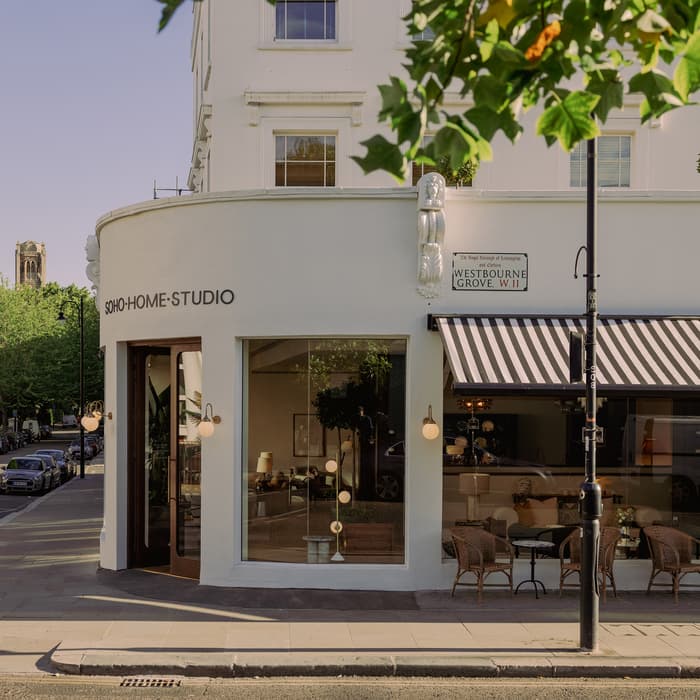 Street corner with Soho Home Studio facade featuring large windows, striped awning, and outdoor seating. Sign on building reads “The Royal Borough of Kensington and Chelsea, Westbourne Grove, W.11.” Tree-lined street and parked cars.