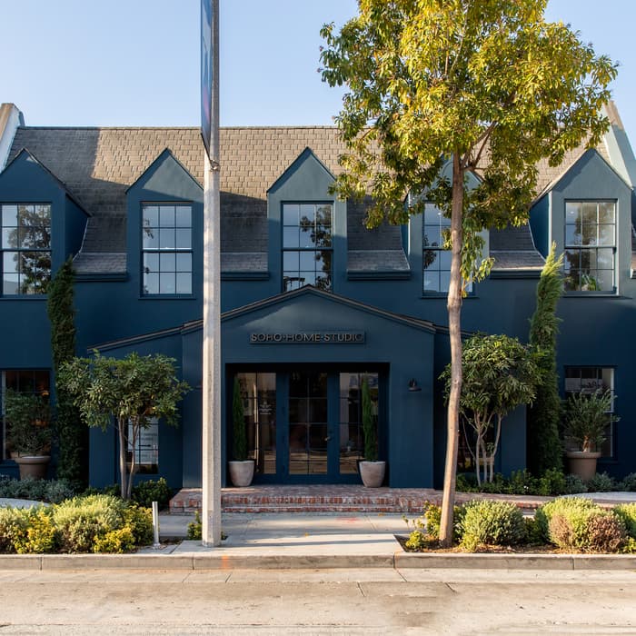 Building labeled "SOHO HOME STUDIO" with large blue facade, sloped roof, tall windows, and surrounding greenery, including trees and shrubs, located on a sunny street.