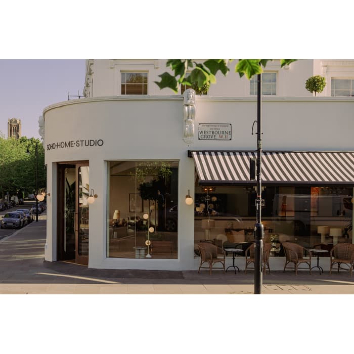 A corner building labeled "SOHO HOME STUDIO," with striped awning, large windows, outdoor seating, and street signs reading "Westbourne Grove W.11," on a sunny street lined with cars and trees.