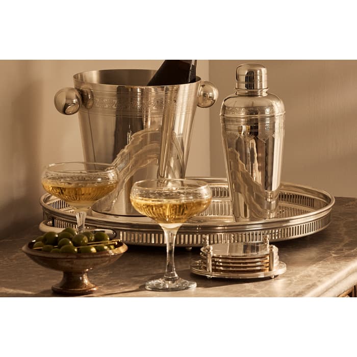 A bottle rests in an ice bucket beside a cocktail shaker, two filled glasses, and a dish of green olives on a marble counter in a warmly lit room.