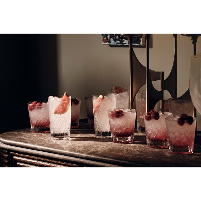 Eight iced cocktails containing berries or fruit garnish rest on a marble countertop beside a golden candlestick in a softly lit room with a framed artwork on the wall.