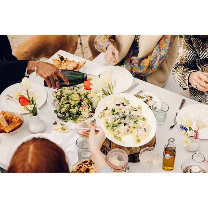 People are enjoying an outdoor meal at a white table, adorned with salads, bread, and drinks. A man pours champagne, and a dog is in the background near a garden.