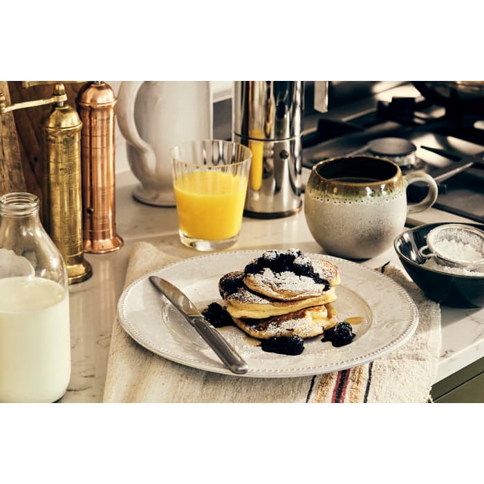 Pancakes topped with blueberries and powdered sugar, placed on a white plate with a knife, surrounded by a bottle of milk, a mug, orange juice, and cooking utensils on a kitchen counter.
