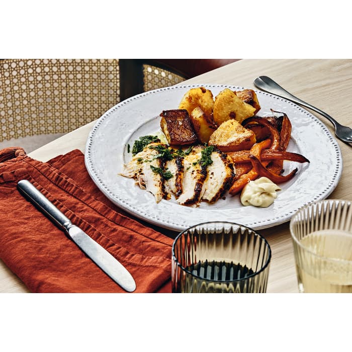 Dinner plate with sliced chicken, roast potatoes, and carrots. Nearby, a glass of wine, a black-handled knife on a red napkin, and wooden chair at a wooden dining table.