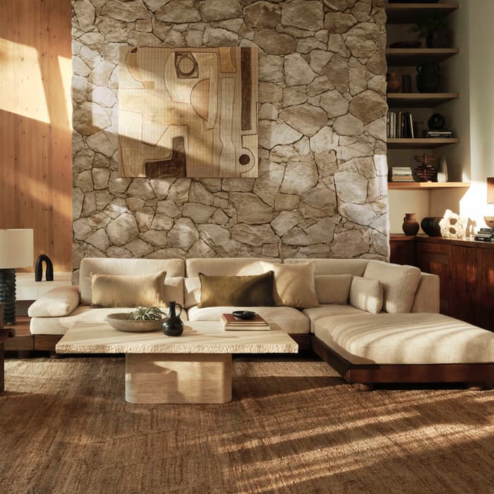 A beige sectional sofa and a wooden armchair sit in a cozy living room with a stone wall, large plant, abstract art, and wooden shelves filled with decor items.