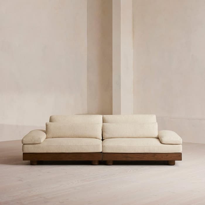 A beige, two-seater sofa with wooden base sits against a pale, minimalist wall in a spacious, softly lit room with light wooden flooring.