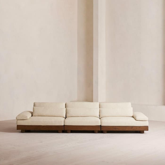 A beige three-seater sofa with wooden legs is positioned against a plain, light-colored wall in a minimalistic, empty room with a wooden floor.