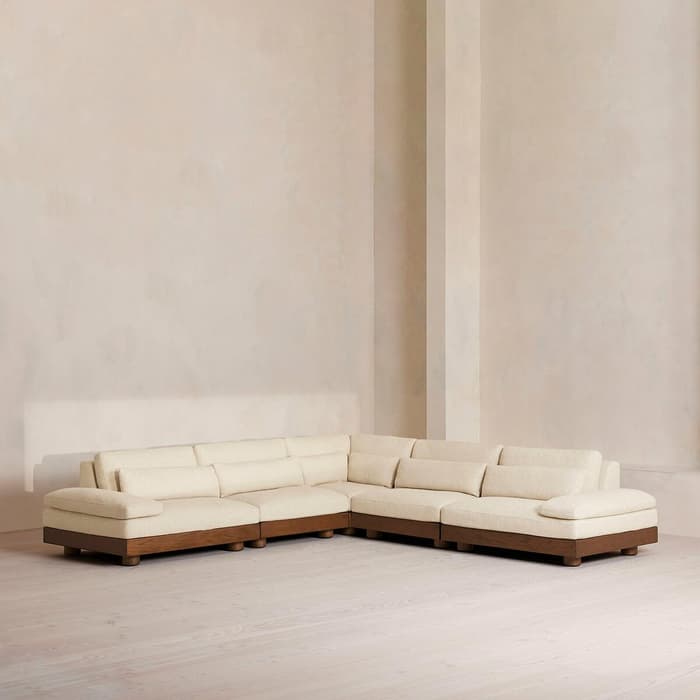 A cream-colored sectional sofa with wooden legs sits in the corner of a minimalist room with light taupe walls and pale wooden flooring.