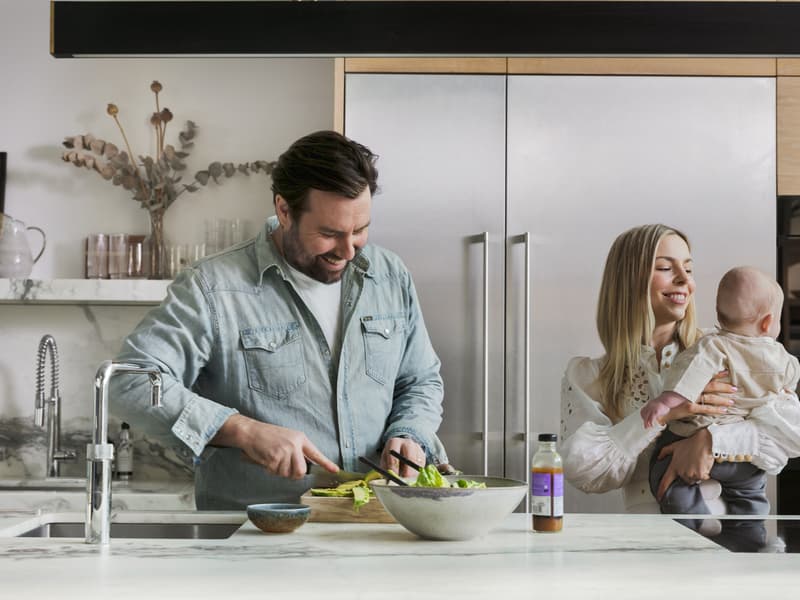 Man preparing a salad in a modern kitchen while smiling, with a woman holding a baby nearby. The kitchen features a marble countertop, sink, and stainless steel appliances.