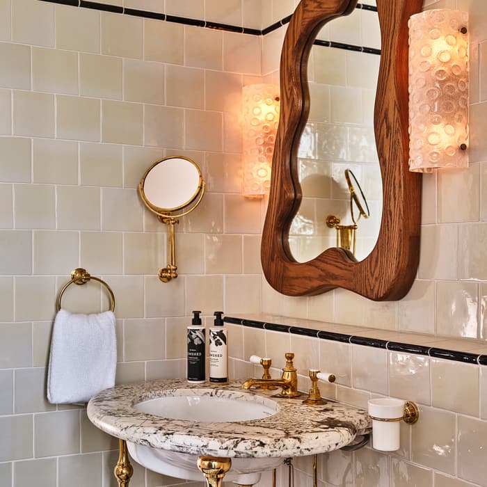 A marble-topped sink with golden fixtures and white tile walls features a wooden-framed mirror, two textured light sconces, a towel ring with a white towel, and two soap dispensers.
