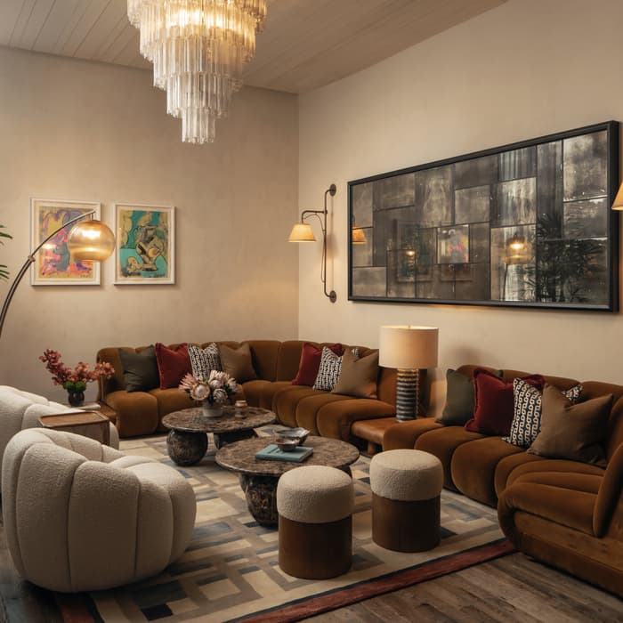 A cozy living room with a brown sectional sofa, cushioned stools, round marble tables adorned with flowers, and framed artwork on beige walls, under a large, cascading chandelier.