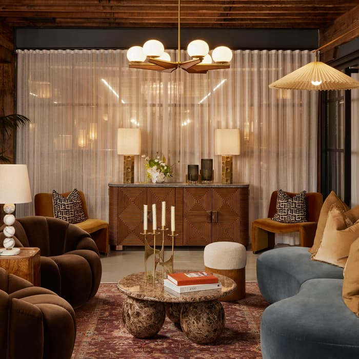 A cozy living room with plush chairs and a curvy sofa arranged around a marble coffee table with a candelabra and books, accented by tall plants and soft, warm lighting.