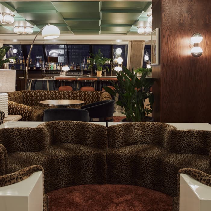 A leopard print sofa sits in a stylish lounge area with plants, tables, and lamps, against the backdrop of a bar with stools and various bottles in the background.